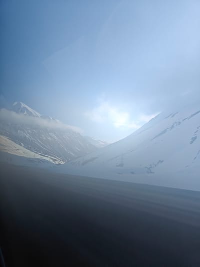 The image depicts a snowy mountain landscape in Ganisi, Georgia. The foreground is a dark, indistinct surface, likely part of a road or vehicle interior. Beyond this, snow-covered mountains rise into a hazy, bright sky. Power lines are visible traversing the slopes. A cloud bank partially obscures the peak of the left mountain. The overall impression is one of a cold, high-altitude environment, seen from a low vantage point.