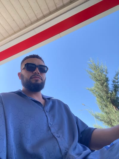 A low-angle, medium shot captures a man wearing large, black sunglasses and a patterned, dark blue button-down shirt. He has dark hair and a beard. The man is outdoors, with a clear blue sky dominating the background. A portion of a structure with a white ceiling and a red stripe is visible at the top of the frame. To the right of the man, a green coniferous tree extends upwards. A street lamp is also visible in the background. The image appears to have been taken on a sunny day. The man is positioned centrally in the frame, with the focus on his face and upper torso. There are no visible interactions with other people. The overall impression is of a casual outdoor portrait.