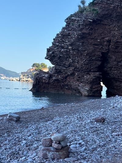 This is a photograph of a rocky beach in Sveti Stefan, Montenegro. In the foreground, the beach is covered in small, smooth pebbles. A small cairn of stones is stacked on the right side of the foreground. In the middle ground, calm, clear blue water meets the shore. A large, dark rock formation with a natural archway dominates the right side of the frame, partially obscuring the view of a small island in the distance. This island, visible in the background, is covered with trees and appears to have buildings with red-tiled roofs. The island is connected to the mainland by a narrow causeway. The sky above is a clear, pale blue. The overall scene depicts a serene coastal landscape.