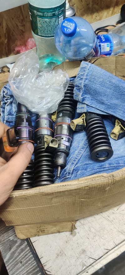 In Rizunenkove, Ukraine, a person's hand is holding a single diesel injector in a cardboard box. The box contains several other diesel injectors, some with springs attached. The injectors are metallic, with visible rings of purple and orange. Jeans fabric is visible underneath and around the injectors, suggesting they may be stored or transported in clothing. A clear plastic bag with a white, crumpled interior is also present in the box, along with two plastic bottles, one clear and one blue. The blue bottle has a cap with Cyrillic text that reads "ПРИРОДНЕ ДЖЕРЕЛО" (Natural Source). The background is indistinct, appearing to be a wooden surface and possibly other items on a shelf. The scene is lit with a bright light source, casting some shadows.