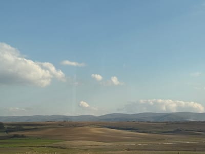 The image displays a rural landscape scene in the area of Baiachevo, Bulgaria. The upper portion of the frame features a light blue sky with scattered white cumulus clouds. A distant, hazy range of undulating hills or low mountains, appearing blue-grey and partially tree-covered, defines the horizon. The midground is characterized by rolling agricultural fields, exhibiting diverse shades of brown from recently tilled soil and green from vegetation. A subtle, light-colored track or path is visible traversing a field horizontally in the lower right section of the midground. A small cluster of structures is faintly discernible on a distant elevation within the central midground. The foreground consists of a narrow strip of mixed green and brown uncultivated ground. No individuals, animals, or direct human activity are present in the scene. The landscape is illuminated by natural daylight.