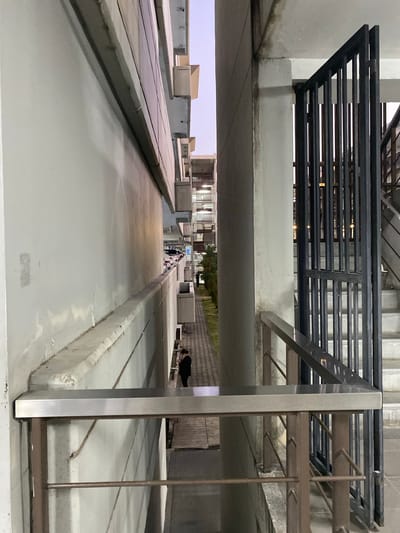 The image shows an outdoor architectural structure in Tambon Sa Khu, Thailand. The scene is dominated by concrete structures arranged in linear pathways, with low concrete walls bordering them. The pathways appear to be at different elevations. In the background, figures of people are visible walking between the structures. Overhead, metal railings and concrete overhangs create a layered, semi-enclosed space. The lighting is ambient and somewhat dim.