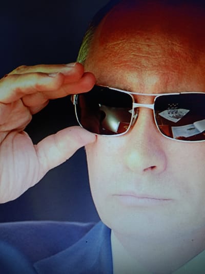 The image shows a close-up of a person wearing aviator sunglasses and a blue suit jacket. The person's right hand is raised to their forehead, as if saluting or shielding their eyes from the sun. The sunglasses reflect a room with a table and what appear to be documents or papers. The lighting suggests an indoor environment. The image does not contain any information that would indicate its location in the city of Молодёжный, Kazakhstan.