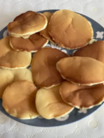A plate of pancakes is shown from a top-down view. The pancakes are golden brown with some lighter golden brown edges. They are piled on top of each other on a dark blue plate with a white pattern. The plate is on a white, textured surface, likely a tablecloth. The image appears to be taken in a home setting in Sumgayit, Azerbaijan.