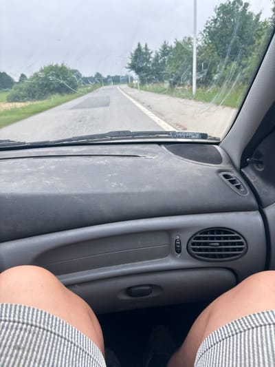 The image is a point-of-view shot from inside a car, looking forward through the windshield. The windshield is dirty with streaks and smudges. A road with painted lines is visible, stretching into the distance with trees and foliage on either side. The sky is overcast. The car's dashboard, including the passenger side air vent and part of the glove compartment, is in the foreground. The lower part of the image shows a person's legs, wearing striped shorts, sitting in the passenger seat. The scene is consistent with driving on a road near Grębanin, Poland.