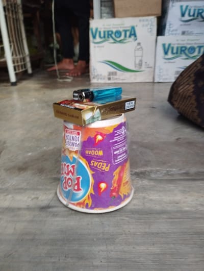 The image captures a ground-level view of several items in what appears to be an indoor or semi-outdoor setting in Kecamatan Panji, Indonesia. In the immediate foreground, an inverted cup of instant noodles, branded "Pop Mie", serves as a base. Stacked on top of the cup is a golden-colored pack of cigarettes, identifiable as "Gudang Garam", with a blue disposable lighter resting on the cigarette pack.
The "Pop Mie" cup features a purple and orange design with red chili peppers, indicating a spicy flavor, and includes text such as "PEDAS" (Spicy), "WOOAH", and "PANGSIT JONTOR". The "Gudang Garam" cigarette pack prominently displays "2.0 MG TAR" and "0.2 MG NICOTINE".
In the background, out of focus, are the bare feet and lower legs, clad in black pants, of a person standing. Behind them, several large white cardboard boxes are visible, labeled "VUROTA" with a stylized image of a water bottle and a green wave logo, suggesting they contain bottled water. To the far left, a portion of a white, possibly metal, frame or shelving unit is discernible. To the right, a textured brown fabric object can be seen.
The scene depicts a static arrangement of everyday consumer items. No specific activity is actively taking place other than the person standing in the background. The general lighting suggests it is daytime. The presence of Indonesian brand names like "Pop Mie" and "Gudang Garam", along with "VUROTA" water, aligns with the specified location context. There are no discernible emotions or specific weather conditions evident.