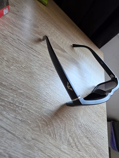 A pair of black sunglasses with a silver emblem on the left arm and "M-2686" printed on the right arm rests on a light brown, wood-grain patterned surface. The sunglasses are positioned with the arms extended and the lenses facing downwards. The surface appears to be a table or desk. In the background, a dark, striped material is visible, possibly a curtain or upholstery. To the far right, a portion of a dark object with a purple fabric element is slightly out of focus. The scene is lit from the left. The location in Mijas, Spain, is not discernible from the visual information in the image.