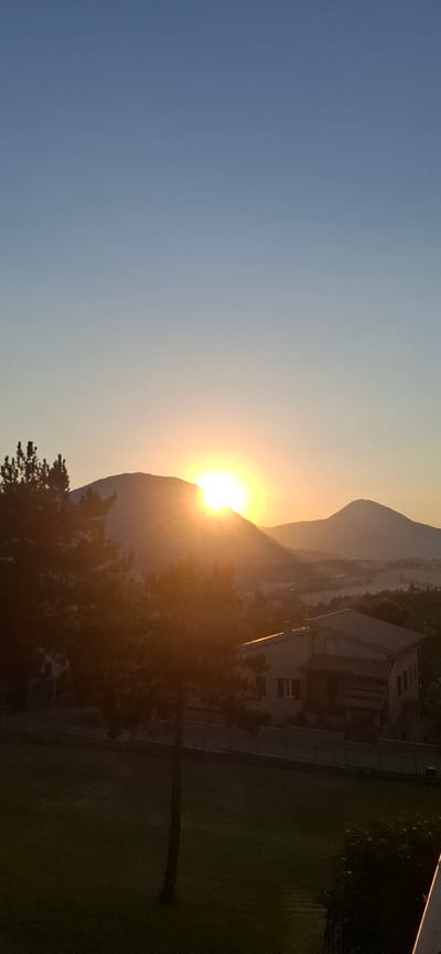 The image captures a panoramic view of the landscape surrounding Camerino, Italy, at sunrise. The sun is emerging from behind a large mountain on the left, casting a bright, warm glow across the sky and illuminating the foothills. In the mid-ground, a residential building with a terracotta roof is visible, nestled amongst trees. The foreground features a lush green lawn with a slender tree trunk standing prominently. The sky transitions from a pale blue at the top to a soft, warm hue near the horizon, indicative of the early morning light. The overall scene conveys a sense of tranquility and natural beauty in the Italian countryside.