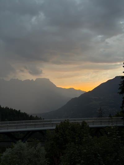 The image depicts a mountainous landscape with a bridge in the foreground. The sky is overcast with dramatic clouds, but a sliver of warm, orange light is visible on the horizon, suggesting either sunrise or sunset. The mountains are layered and shrouded in mist, creating a sense of depth. In the middle ground, a modern-looking bridge with railings spans across a valley, connecting two forested slopes. The foreground is dominated by dense, dark green foliage, including trees and bushes. The overall atmosphere is moody and serene, with the muted colors of the sky and the dark tones of the landscape conveying a sense of natural beauty. The presence of the bridge indicates human infrastructure within the natural environment. Based on the architecture of the bridge and the style of the surrounding buildings visible in the distance on the mountainside, this scene is likely located in the vicinity of Saint-Gervais-les-Bains, France, a town known for its mountainous terrain and ski resorts.