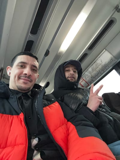 Two men on a public transport vehicle pose for a selfie. The man closest to the camera wears a jacket with an orange shoulder section, while the other man wears a darker jacket and cap. One of them gestures with his fingers. A subway map is seen.