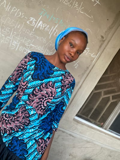 A woman wearing a blue and pink patterned long-sleeved top and a blue headscarf poses in front of a wall. The wall has white chalk writing on it and a window frame in the background. Her expression is a soft pout. The angle suggests the photo was taken from slightly above. The lighting is bright and natural.