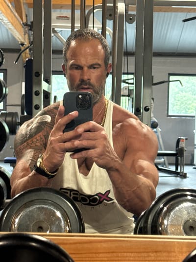 A man with short, dark hair and a beard is taking a selfie in a gym. He is wearing a white tank top and a gold chain necklace. He has tattoos on his left arm and is wearing a watch on his left wrist. In the foreground, there are weight plates. In the background, there is gym equipment and a window with trees visible outside. The image appears to have been taken in Jemelle, Belgium.