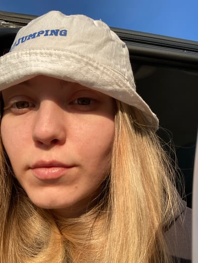 A close-up portrait of a fair-skinned person with long blonde hair is wearing a white bucket hat with the word "JUMPING" printed vertically in blue on the left side. The individual is positioned indoors near a window or opening with a view of the sky. The sky appears blue. The lighting is strong, creating shadows across their face. The person is facing the camera with a neutral expression. The background shows a partial view of a dark, possibly black, interior frame.