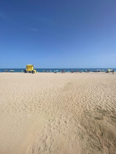 The image depicts a wide, sandy beach on a clear, sunny day. The foreground is dominated by the textured surface of the sand, showing ripples from the wind and scattered footprints. In the mid-ground, a line of beachgoers is visible, some sitting under colorful umbrellas, others standing or walking along the shore. A bright yellow lifeguard tower stands out prominently on the left side of the mid-ground, with a dark green trash receptacle next to it. The background features the calm, blue water of the Mediterranean Sea, meeting the clear blue sky at the horizon. The scene appears to be located in Vilassar de Mar, Spain, as indicated by the provided context, suggesting a typical summer day at this coastal town.