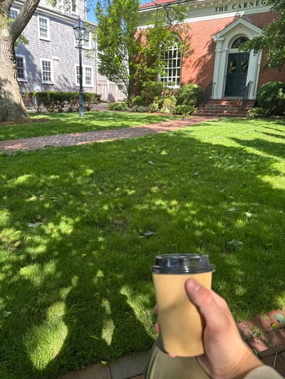 A person's hand holds a disposable coffee cup in front of a manicured lawn and brick walkway, with "THE CARNEG" visible on a brick building on the left. A grey shingled building and a green street lamp are visible behind the trees in the distance. The scene is bathed in dappled sunlight, creating shadows across the grass. The image was taken in Edgartown, United States of America, specifically near the Carnegie museum.