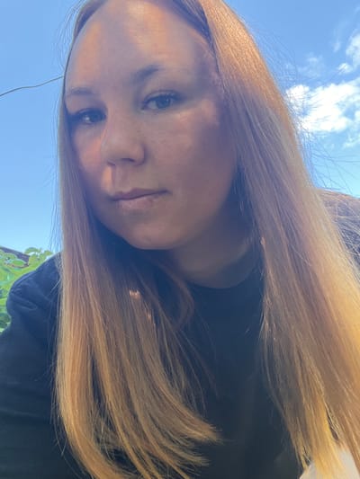 A close-up, low-angle selfie shows a young woman with long, blonde hair and fair skin. She is looking directly at the camera with a neutral expression. Her hair is falling forward, partially obscuring her face. She is wearing a dark t-shirt. The background is a bright blue sky with some wispy white clouds. There are some green leaves visible in the lower left corner of the image, suggesting an outdoor setting. The image was taken in Arzh, Russia.