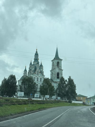 An overcast, cloudy day in Nizhnyaya Sinyachikha, Russia, features a prominent, light blue and white church with multiple domed towers and a tall, slender steeple. The church is partially obscured by lush green trees. In the foreground, a paved road curves to the right, with a grassy embankment running alongside it. Several informational signs are visible on the embankment. To the right of the church, in the background, are several brightly colored, traditional Russian houses. Power lines are visible against the sky.