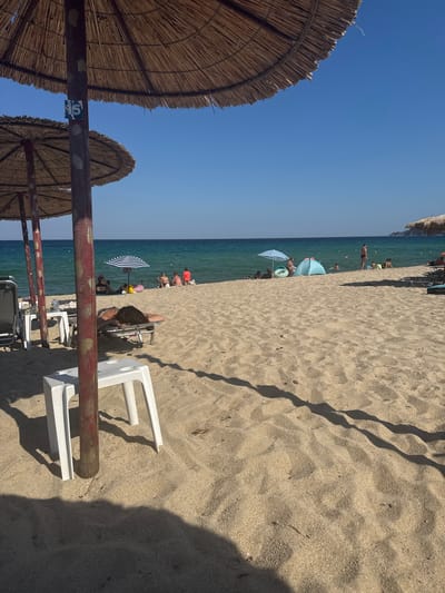 The image depicts a sunny beach scene in Toroni, Municipal Unit of Sithonia, Greece. In the foreground, the sandy beach is dotted with shadows cast by thatched umbrellas, which provide shade for lounge chairs and small white plastic stools. One person is visible lying on a lounge chair under an umbrella, facing away from the camera. Further back, near the turquoise water, several people are gathered, some sitting on the sand, others standing. A striped umbrella is set up, and a light blue tent is visible. People are wading in the shallow water and swimming in the sea. The sky is clear and blue. The overall impression is that of a tranquil summer day at the beach.