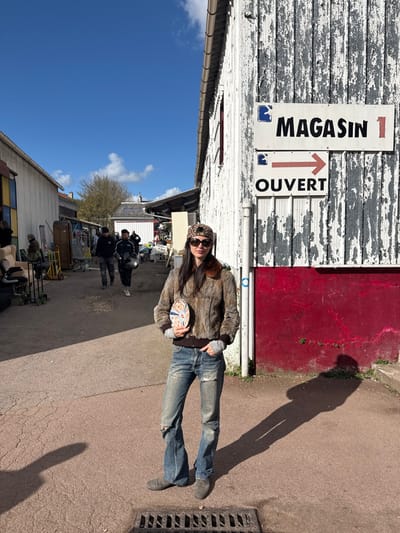 The image depicts an outdoor marketplace or flea market area in Neuilly-Plaisance, France, under a clear blue sky with scattered white clouds. In the foreground, a female subject stands facing the viewer, slightly off-center to the left. She has long dark hair, wears dark sunglasses, a brown and black leopard-print baseball cap, a brown faux fur bomber jacket with a dark brown ribbed collar and cuffs, grey long-sleeved undershirt, distressed blue flared jeans, and grey shoes. She holds a circular decorative plate with a colorful design in her left hand at waist level.
To her right, a weathered white corrugated building with peeling paint features a vibrant red painted base and a white vertical pipe. Two signs are affixed to this building: an upper sign reads "MAGASIN 1" (Store 1) with a blue emblem, and a lower sign reads "OUVERT" (Open) with a red right-pointing arrow.
Behind the female subject, a paved reddish-brown ground with visible cracks and a metal drain grate in the lower foreground extends into an alleyway. Further down the alley, two male figures walk away from the viewer; one is in a dark jacket, and the other wears a dark jacket and holds a black motorcycle helmet. Various market stalls, furniture, and miscellaneous objects are visible along both sides of the alleyway in the mid-ground and background. Another building, similar in style but less detailed, stands to the left. Long shadows are cast on the ground from the right, indicating strong sunlight.