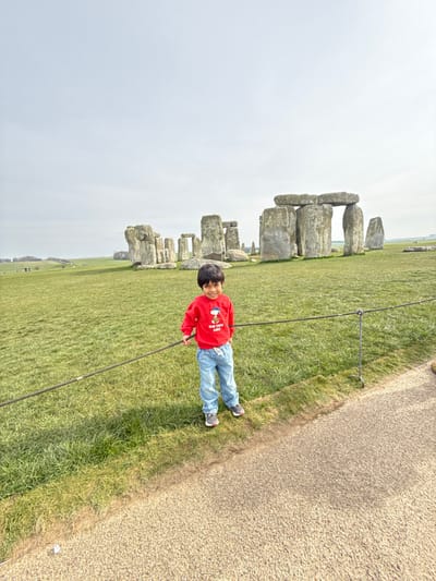 The image depicts an outdoor scene at Stonehenge, located near Amesbury, Wiltshire, United Kingdom. In the foreground, a young child stands on a grassy area behind a dark rope barrier that separates it from a gravel path visible on the right. The child, appearing male with dark hair, faces the camera and smiles, wearing a red long-sleeved shirt with a graphic of Snoopy and Woodstock, light blue jeans, and grey athletic shoes. His left hand is positioned near the rope barrier. In the mid-ground, the ancient megalithic structure of Stonehenge is visible, consisting of large sarsen stones arranged in a circular formation, with several vertical stones topped by horizontal lintel stones. The surrounding environment consists of expansive green fields under a light grey, overcast sky. Small, indistinct figures of other individuals are visible in the far distance on the left side of the frame.