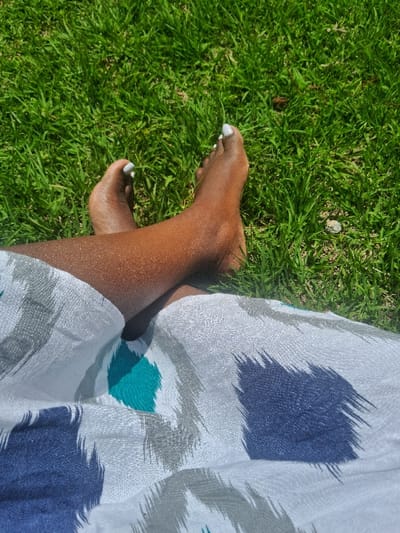 The image depicts a close-up, high-angle view of a person's bare lower legs and feet resting on green grass in Muthambi ward, Kenya. The person's skin tone is dark brown, and the feet are crossed at the ankles, with both visible. Toenails on both feet are painted a solid white. The person is wearing a garment over their lap and upper legs, featuring an abstract pattern of large, textured shapes in dark blue, teal, and gray against a white background. The grass beneath the feet is vibrant green and appears well-lit by natural sunlight, indicating an outdoor setting. No other individuals or objects are discernible.