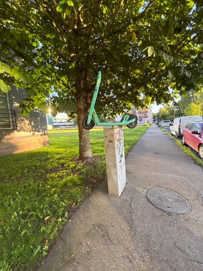 This media file depicts an outdoor scene in Tartu, Estonia. The primary subjects are a green electric scooter leaning against a tree, a sidewalk, a grassy area, and parked vehicles. The electric scooter is a prominent object, positioned in the mid-ground, with its front wheel visible. The tree, with its dense green foliage, is on the left side of the frame and partially obscures the scooter.
The setting appears to be a residential or urban area, with a sidewalk running alongside a street where several cars and vans are parked. The vehicles include a red car, a white van, and other cars further down the street. The sidewalk is made of asphalt and shows some cracks, with a manhole cover visible on it. The grassy area next to the sidewalk is well-maintained and appears to be a lawn. In the background, there are buildings, suggesting a developed area.
The time of day appears to be late afternoon or early evening, as indicated by the warm, angled sunlight casting shadows and illuminating the scene. The weather seems pleasant, with clear skies, as suggested by the bright sunlight and the absence of any signs of rain or overcast conditions.
There is some visible text, most notably graffiti and stickers on a metallic structure located near the scooter and the tree. The graffiti is colorful and abstract, adding a touch of urban character to the scene. The electric scooter itself has some branding, but it is not clearly legible in the image.
No people are visible in the media file. The overall mood is calm and peaceful, capturing a typical everyday scene in an urban environment. The image is taken from a low angle, emphasizing the tree and the scooter.