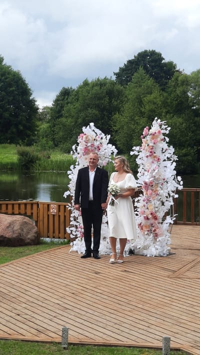 A wedding ceremony is taking place outdoors on a wooden deck in Жодзішкаўскі сельскі Савет, Belarus. A man in a black suit and a woman in a white dress are standing hand-in-hand in front of a floral wedding arch. The arch is decorated with white and pink flowers and branches. Behind them, a body of water is visible, with lush green trees lining the banks. The sky is overcast with clouds. The wooden deck is situated on a grassy area, with a large rock and a wooden fence to the left of the couple.