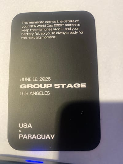This is a close-up, sideways shot of a black card, likely a ticket or souvenir for a FIFA World Cup 2026 match. The card is held in a horizontal orientation, with the text running vertically. The background is a plain white surface. The text on the card describes the item as a "memento" that carries the details of a "FIFA World Cup 2026™ match" and is designed to help "keep the memories vivid" and ensure the "battery full."
Prominently displayed are the details of a specific match: "JUNE 12, 2026," "GROUP STAGE," and the location "LOS ANGELES." Below this, it lists the participating teams: "USA v PARAGUAY." The text is predominantly white and sans-serif, with "GROUP STAGE" being the largest and boldest. The overall aesthetic is sleek and modern. The lighting suggests an indoor setting, possibly with a slight overhead light source. There are no people or clear environmental cues beyond the card itself, making it difficult to determine a specific time of day or weather. The context provided places the event in Inglewood, United States of America, aligning with the "Los Angeles" location mentioned.