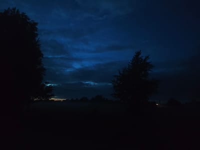 The image depicts a dark, rural landscape at dusk or night, likely within the Яглевіцкі сельскі Савет region of Belarus. A large, silhouetted tree dominates the left side of the frame. In the mid-ground, the faint outlines of trees and possibly structures are visible against a dark, hazy horizon. The sky is a deep, dark blue with some wispy clouds that catch the fading light. The overall atmosphere is somber and quiet. There are no people or animals visible in the scene. The lighting suggests it is either twilight or night, with minimal artificial light sources, save for a faint distant illumination that could be a headlight or a solitary lamp. The foreground is entirely obscured by darkness.