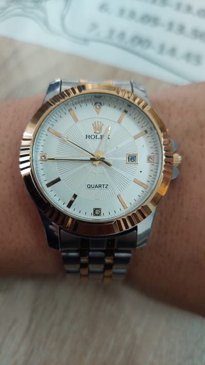 The image shows a close-up of a two-tone (gold and silver) wristwatch on a person's wrist. The watch face is white with gold-colored hour markers, and the word "QUARTZ" is printed on the right side of the face. The date is displayed as "16" in a small window. The band is two-tone and metal. The background is a light brown surface, possibly wood. The location is not discernible from the image.