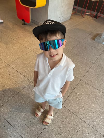 A young boy wearing a black baseball cap and futuristic, mirrored sunglasses smiles at the camera. He is dressed in a white short-sleeved polo shirt and light blue shorts. The boy is wearing light-colored sandals on his feet. He appears to be standing on a tiled floor, possibly indoors. To the left of the frame, a red and yellow bench is visible. The lighting suggests it could be daytime, and the indoor setting implies it is not weather-dependent. There is some visible text on the boy's cap, which is partially obscured, but "REAL" is legible. The overall impression is one of a cheerful, stylish child posing for a photo in a public or recreational space in Katowice, Poland.