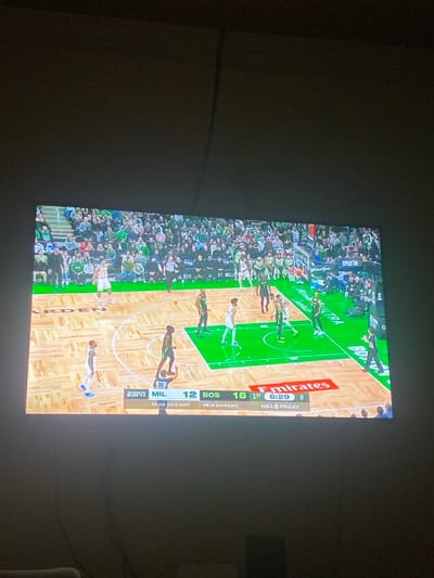 A large flat-screen television displays an NBA basketball game in progress, with the image rotated 90 degrees clockwise. The screen shows several basketball players on a wooden court, dressed in green and white uniforms. A crowd of spectators is visible in the stands behind the court. The setting appears to be an indoor basketball arena, viewed on a TV in a dimly lit room, likely a home environment in Lakka, Sierra Leone.

A score bug overlay is prominently featured on the right side of the screen. Visible text includes "MIL 12" and "BOS 16," indicating the Milwaukee Bucks are trailing the Boston Celtics. The game is in the "1st" quarter with "6:29" minutes remaining. Other text includes "NBA FRIDAY," the "ESPN" channel logo, and sponsor logos such as "Emirates" and "ULTRA." Below the scores, team records are partially visible: "11-10 (EAST)" for Milwaukee and "10-4 ON LAST" for Boston. The text "Pick Em" is also visible in the top left corner of the screen. The overall lighting of the room is dim, highlighting the bright screen displaying the game.