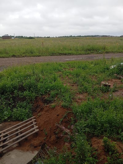 The image depicts an overcast outdoor scene in the village of Azmukino, Russia. In the foreground, there is a pile of reddish-brown soil with green vegetation growing around it. To the left, a wooden pallet lies on a concrete surface. A dirt road with some puddles runs horizontally across the middle ground. Beyond the road, a large field of green grass and wildflowers stretches towards the horizon. In the distance, several houses and trees are visible, along with utility poles and wires. The sky is heavily clouded.