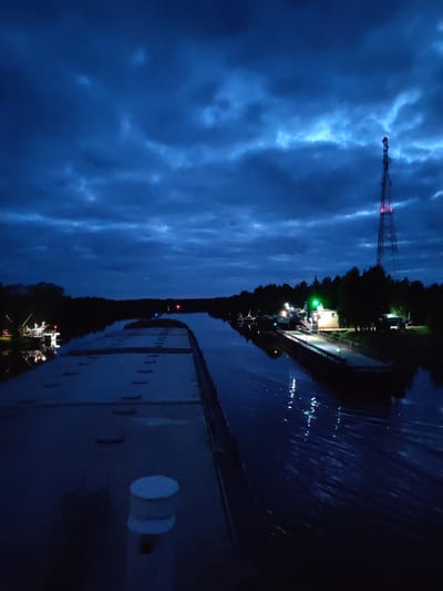 The image depicts a nighttime scene at Annensky Most, Russia. A canal flows through the scene, reflecting lights from surrounding structures. A barge occupies the foreground, its deck featuring hatches and a bollard. On the right, a small harbor is illuminated with lights. A communication tower stands in the background, its red and white pattern visible against the dark sky. The sky is filled with dark, layered clouds. The overall illumination is low.