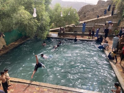 The image displays an outdoor recreational bathing area, specifically a rectangular pool, located in Killi Malik Sher Mohammad, Pakistan. The pool's clear, rippling water occupies the foreground and mid-ground, bordered by reddish-brown paved edges. Lush green trees, resembling willows, line the left side of the pool, with several white, bulb-shaped lights suspended on wires overhead. To the right and in the background, a terraced, rocky hillside rises, constructed with stone retaining walls forming steps and platforms. Beyond the hillside, barren mountains are visible under a bright sky. Approximately 20 to 30 males of various ages, from young children to adults, are present in the scene. Several individuals are actively in the pool, with one male captured mid-dive, headfirst into the water, his legs extended vertically above the surface. Other individuals are swimming, standing in the water, or observing from the paved edges and the elevated stone terraces. Some clothing items are draped over the rocky structures on the hillside, likely for drying. Attire ranges from swimwear and casual clothing to traditional shalwar kameez.