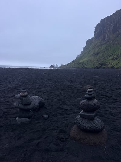 This image captures a scene at the black sand beach in Vik, Iceland. The ground is covered in black sand. In the foreground, two stacks of rocks are arranged in the style of rock cairns. In the background, there are cliffs covered in green vegetation and the ocean. The sky is overcast and gray.