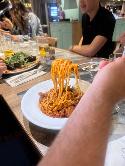 A person is twirling spaghetti onto a fork from a white plate of spaghetti with sauce. In the background, people are seated at a table, and there are glasses and food, including a pizza. The setting appears to be a restaurant, given the casual attire of the diners and the presence of multiple food items and tableware. The precise location in Saint Paul's Bay, Malta cannot be determined from the image alone.
