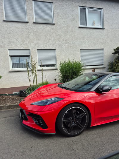 A bright red C8 Chevrolet Corvette, featuring a contrasting black roof and multi-spoke black wheels with Michelin Pilot Sport tires, is parked on an asphalt street in Traben-Trarbach, Germany. The vehicle is angled slightly towards the left, showcasing its aggressive front fascia and dark LED headlights. Behind the car, a two-story building with a light-colored, stucco-textured facade is partially visible. The upper floor displays two windows with light grey external roller shutters fully closed, and one window partially open to the right, revealing an internal decorative element and a screen. On the lower floor, several windows are visible, all featuring closed light grey external roller shutters. A planting bed, covered with dark brown mulch, lies between the street and the building, containing various green plants including tall, spiky foliage and denser bushes. A concrete curb separates the street from the planted area. The scene is illuminated by natural daylight; no individuals are present.