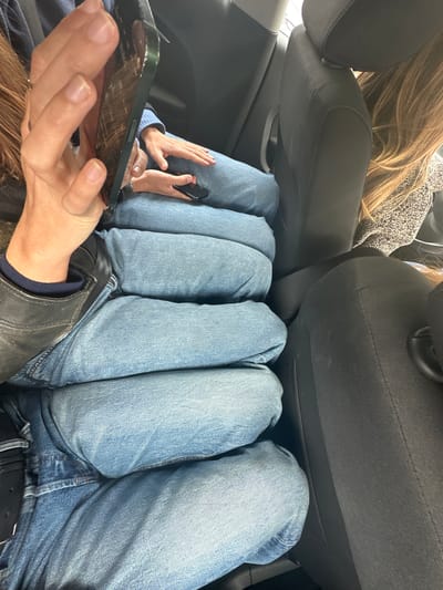 The image shows the interior of a car with four people seated in the back. All four individuals are wearing light blue denim jeans. The legs of the individuals are pressed together, taking up the majority of the available space. One person is holding a smartphone in their hand in the foreground, and another person has a phone in their hand further back. The car seats are dark grey. The location is not discernible from the image.