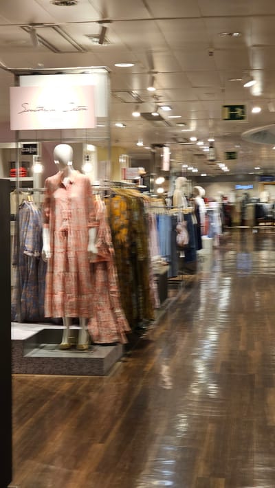 The image shows the interior of a clothing store, likely in Zaragoza, Spain, given the context. The scene is characterized by motion blur, suggesting either a moving camera or a long exposure. In the foreground, a mannequin is dressed in a long, light-pink tiered dress with a patterned design. To the left of the mannequin, another dress with a blue and white pattern is visible on a hanger. To the right, a rack of clothing displays garments with vibrant yellow and green floral patterns, followed by racks of blue and white clothing.

Above the mannequin, a rectangular sign with a pink background displays the text "Southern Sun" in a stylized script. Below this sign, another smaller sign reads "caja." The store has a polished wooden floor that reflects the overhead lighting. Numerous track lights are visible on the ceiling, illuminating the store. In the background, more clothing racks and mannequins are present, along with signage, including a green sign with a white "H" symbol, indicating a medical facility or first aid station. The overall impression is of a well-lit retail environment with a focus on women's fashion.