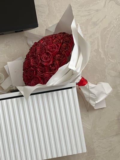 A bouquet of red roses, each adorned with glitter, is presented against a textured, light-colored wall with a marbled pattern. The roses are clustered together, forming a heart shape, and are partially wrapped in white paper, secured with a red ribbon. To the right of the bouquet is a white object with horizontal ribbed detailing, possibly a piece of furniture or an appliance. A black object, likely a box or container, is visible in the lower left corner. The overall scene appears to be indoors, with soft lighting. No people are visible, and there are no clear indications of the time of day or weather conditions. No text is discernable in the image.
