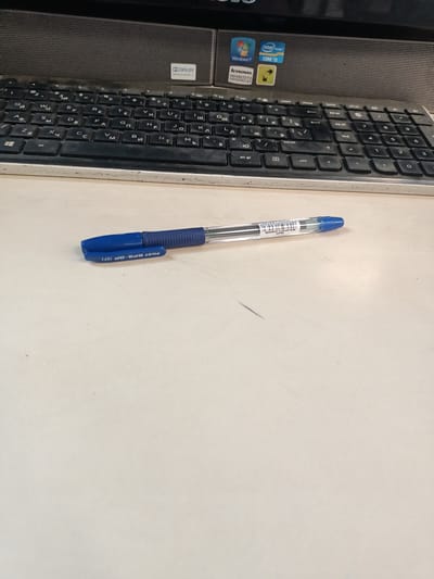 The image displays a blue and silver pen lying on a light-colored surface. The pen has a textured blue grip and a clear barrel with some writing on it. Behind the pen, a portion of a keyboard is visible. The keyboard appears to be black and has Cyrillic characters on the keys, suggesting it is likely a Russian or Eastern European keyboard layout. Above the keyboard, there is a dark gray speaker grille with some stickers on it, including a "Dolby Advanced Audio" logo and stickers for "Windows 7," "Intel Core i5," and "Lenovo Enhanced Experience." The overall scene appears to be a desk area. There are no people visible in the image. The location is stated to be Mikhnevo, Russia.