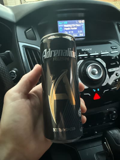 In the interior of a vehicle, likely a Ford Focus, a person's hand holds a can of "Adrenaline Rush" energy drink. The car's dashboard is visible, featuring a Sony audio system with a display showing "Aux < USB TA 19:45 Walking On Cluo.. Heath Hunter Дp. 6/18 3:29 Оскан. Инф". The ambient lighting suggests it is daytime. The vehicle is not in the city of Mayrtup, Russia, as there is no information in the image to support this. The can is black with silver and gold lettering and features a large stylized "A". The hand is pale and the thumb is prominently visible. The car's interior appears to be in good condition, with a steering wheel and gear shift lever also visible.