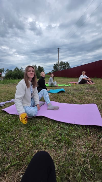 In the city of Kupoyarovo, Russia, a group of four women are seen sitting on yoga mats outdoors on a cloudy day. The woman in the foreground, wearing a white zip-up hoodie and light blue patterned leggings, is holding a yellow mug with a sunflower design. She has light brown hair and is looking towards the camera. Behind her, three other women are seated on yoga mats of various colors (green, blue, and pink). The woman in the black top and dark pants is in the middle ground. Further back, a woman in a light grey top and light pants is seated on a green mat, and another woman in a white top and dark pants is on a pink mat. The women appear to be engaged in a yoga session or a similar outdoor activity. The environment is grassy, with trees and a red fence visible in the background. A telephone pole with wires is also present in the scene. The overall atmosphere suggests a casual, outdoor gathering.