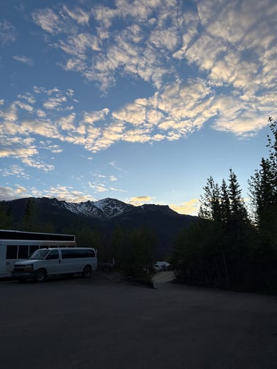 The image depicts a parking lot scene in Denali National Park and Preserve, United States. In the foreground, a dark asphalt surface occupies the bottom portion of the frame. To the left, a white passenger van is parked next to a section of a larger white bus. The van is facing right, and the bus is partially visible. Behind the vehicles, a line of dark green trees forms a dense barrier.

In the midground, a winding asphalt road leads further into the park, with several vehicles parked in the distance, including what appears to be another white van and a red ATV. A scattering of trees is visible along the edge of the road.

The background is dominated by a majestic mountain range. The peaks are rugged and dark, with patches of snow visible on their upper slopes. The sky above is a clear blue, adorned with scattered white and golden clouds, suggesting either sunrise or sunset. The overall lighting indicates a soft, natural light, characteristic of either the early morning or late evening. The scene appears tranquil and natural.