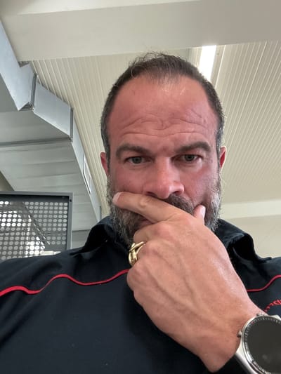 A selfie of a middle-aged man with graying facial hair is oriented at an oblique angle. He is fair-skinned, with visible forehead wrinkles and green eyes. His left index finger is positioned near his mouth as if in thought. He is wearing a black shirt with a red trim along the edge. His left hand has a large gold ring on the ring finger and a watch on his wrist. Behind him is a wall with horizontal textured panels and a piece of metal ventilation equipment. The city of Diekirch, Luxembourg, is not discernable from the image.