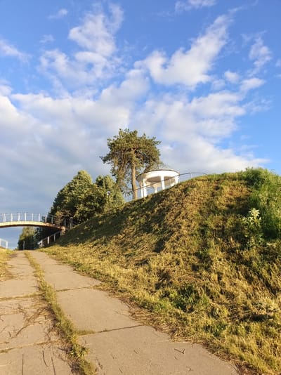 A paved walkway ascends a grassy hill on a sunny day. The walkway, composed of large rectangular concrete slabs, is lined with overgrown grass. At the top of the hill, a white gazebo with a domed roof sits under a large pine tree. A metal railing surrounds the gazebo. In the background, a modern pedestrian bridge arcs across the scene, supported by metal beams and a concrete pier. Lush green trees border the walkway and hill. The sky above is a bright blue, dotted with white, fluffy clouds. The scene appears to be part of a park or public space in the city of Buy, Russia.