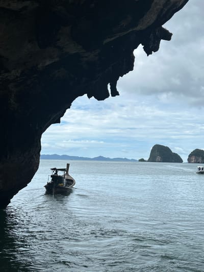 An overcast day at sea in Tambon Krasom, Thailand, features a longtail boat in the foreground, moving away from the viewer. The boat is dark brown with a wooden hull and a prominent rudder. A person is visible in the boat, operating the engine. The water is calm with gentle ripples. A large, dark rock formation overhangs the left side of the frame, casting a shadow on the water and obscuring a portion of the scene. In the distance, several lush green karst islands rise from the sea under a cloudy sky. Another boat, smaller and faster, with two people aboard, is visible to the right, further out at sea. The horizon is lined with a distant mountain range.