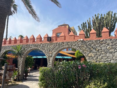 This is a ground-level, eye-level, medium shot of a restaurant's exterior in Tarifa, Spain. The scene is dominated by a rustic stone archway in the foreground, framing a view of an outdoor dining area. Behind the archway, a vibrant coral-pink wall with pointed crenellations runs across the midground. This wall is topped by a distinctive building with a tiled roof, featuring a mix of coral-pink and yellow stucco. Several tall, columnar cacti grow behind the crenellated wall, on the right side of the image.
The outdoor dining area, visible through the archway, includes wicker chairs and tables arranged on a stone patio. Blue and orange awnings provide shade over the seating areas. Lush green bushes and small pink flowers line the bottom edge of the frame, in front of the stone archway. A palm tree trunk is visible on the left side, partially obscuring the upper left corner of the scene. The sky above is a clear blue with wispy white clouds. The overall impression is a charming, Mediterranean-inspired setting, likely a tourist-oriented establishment given its architectural style and landscaping.