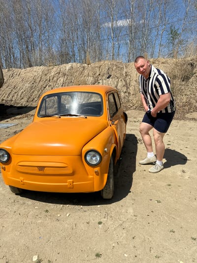 The image depicts a man standing next to a small, orange car. The man is wearing a striped shirt and navy shorts. The car is parked on a dirt surface with sparse vegetation. In the background, there are trees and a blue sky. The image was taken outdoors in Sadovoe, Russia.